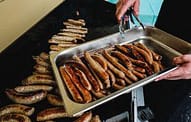 Sizzling sausages cooking on a stovetop grill in a professional kitchen setting.
